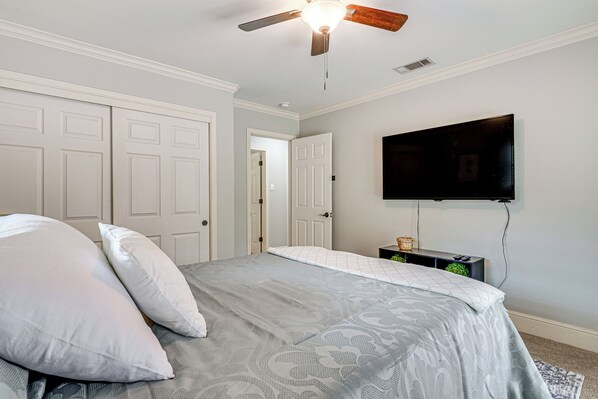A bedroom with a bed, white walls, a ceiling fan, a mounted TV, and a door leading to another room.