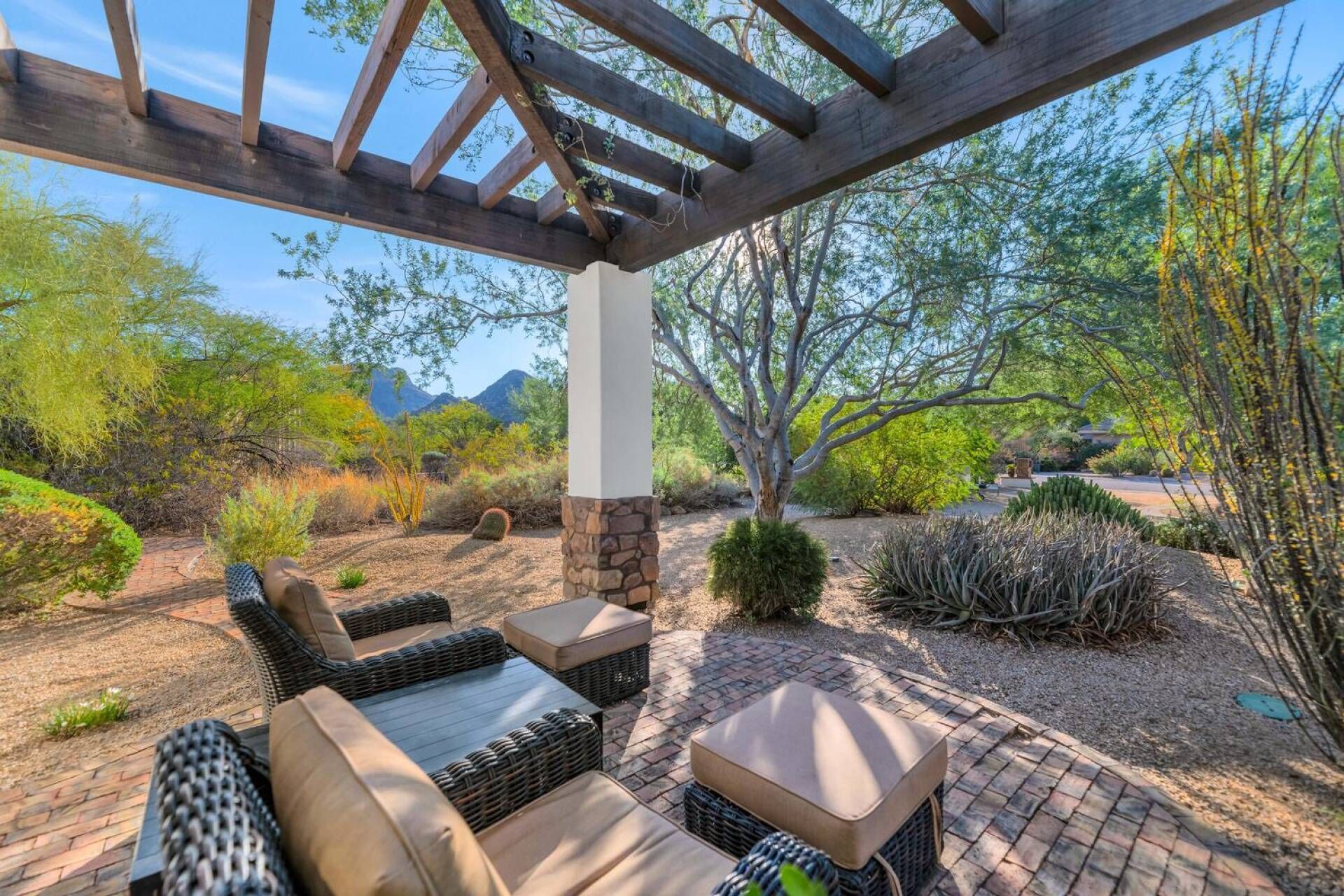A covered outdoor seating area with wicker furniture and stone pillars, surrounded by desert vegetation and mountains in the background.