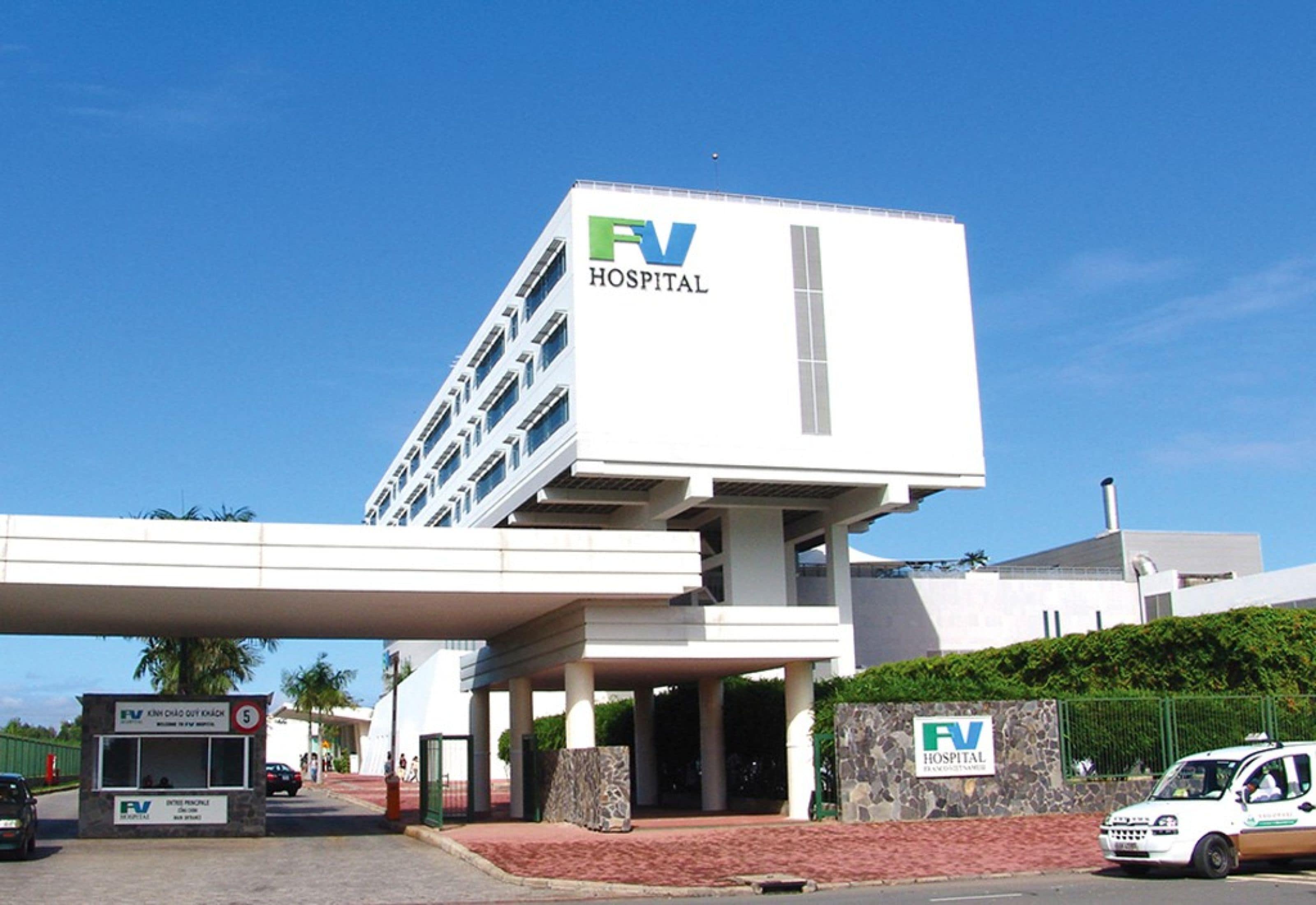 Un bâtiment d'hôpital avec une enseigne bien visible indiquant « Hôpital FV », une entrée couverte et un espace de stationnement où sont garés quelques véhicules.