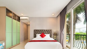A hotel room with a large bed, a grey upholstered headboard, a red pillow, a bedside lamp, and a view of palm trees through the window.