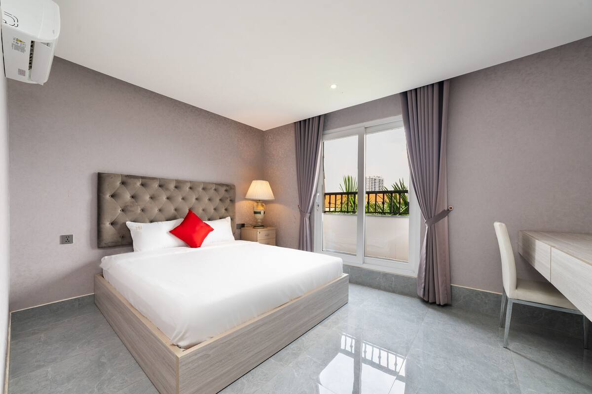 A modern hotel room with a large bed, a bedside table with a lamp, a desk, and a view of a balcony with plants.