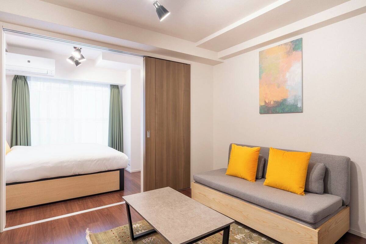 A hotel room with a bed, a sofa with yellow cushions, a small table, and a painting on the wall.