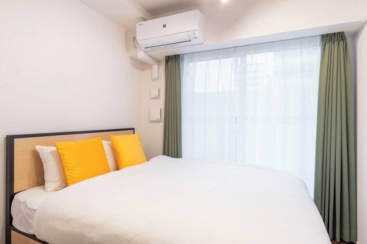 A hotel room with a bed, a window with green curtains, and an air conditioning unit mounted on the wall.