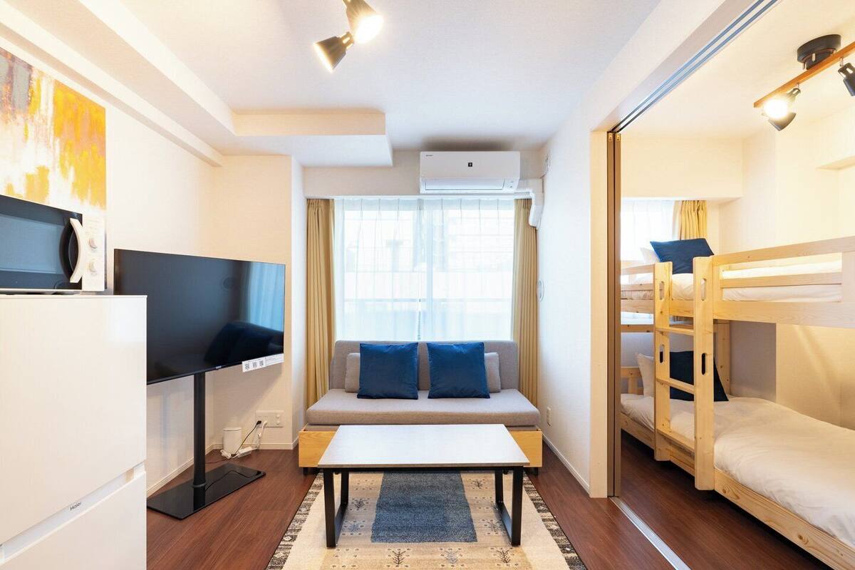 A compact hotel room with a sofa, a TV, a small table, bunk beds, and an air conditioning unit.
