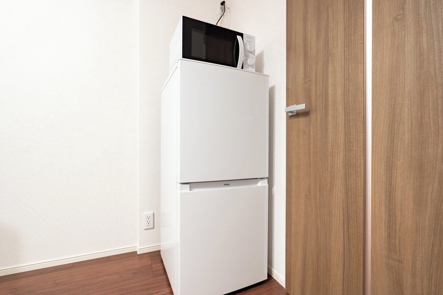 Ein weißer Kühlschrank mit einer Mikrowelle darüber, neben einer Holztür in einem Raum mit weißen Wänden und Holzboden.