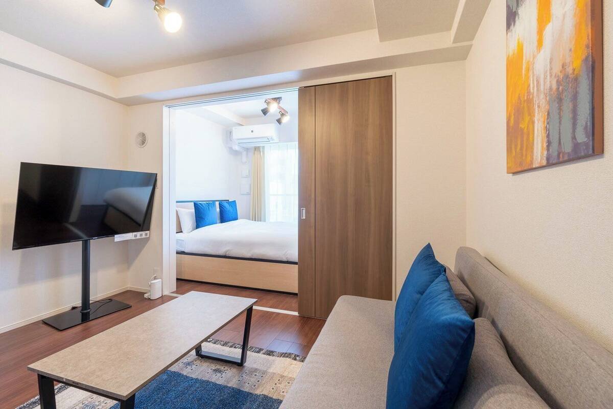 A modern hotel room with a flat-screen TV, a sofa, a bed with blue pillows, and a painting on the wall.