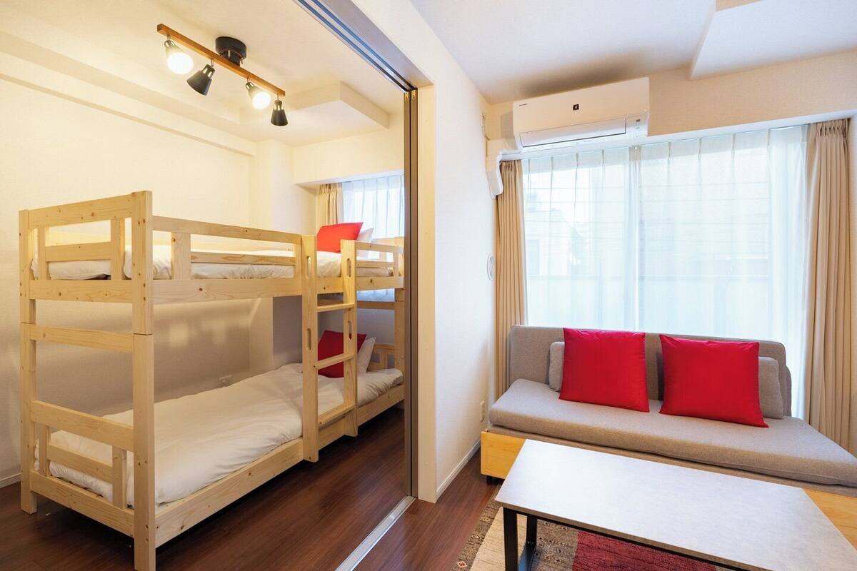 A room with bunk beds, a sofa with red cushions, a small table, and an air conditioning unit.