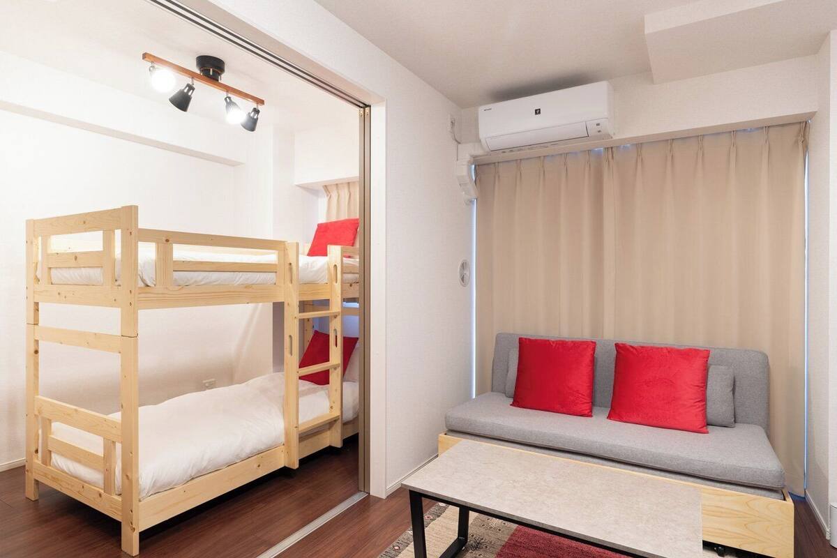 A bunk bed with a desk and a sofa with red cushions in a room with an air conditioner and curtains.