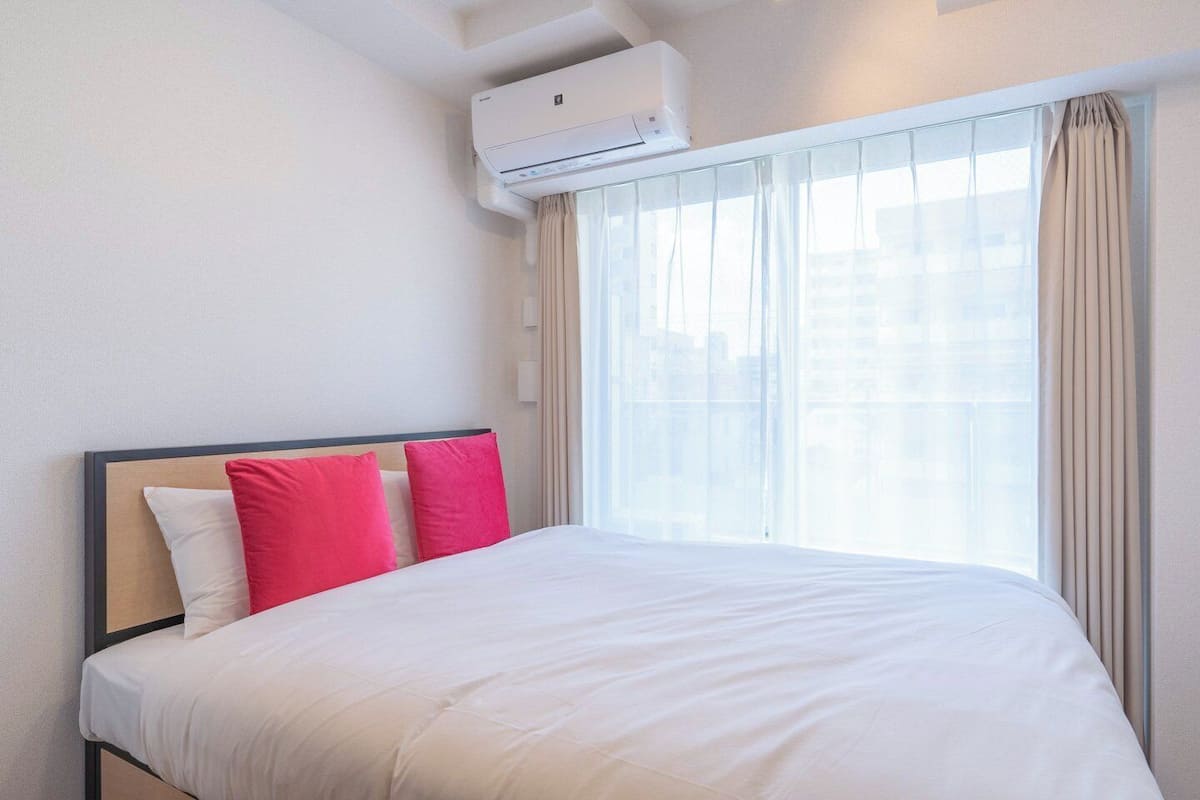 A hotel room with a bed, white bedding, a window with sheer curtains, and an air conditioning unit mounted on the wall.