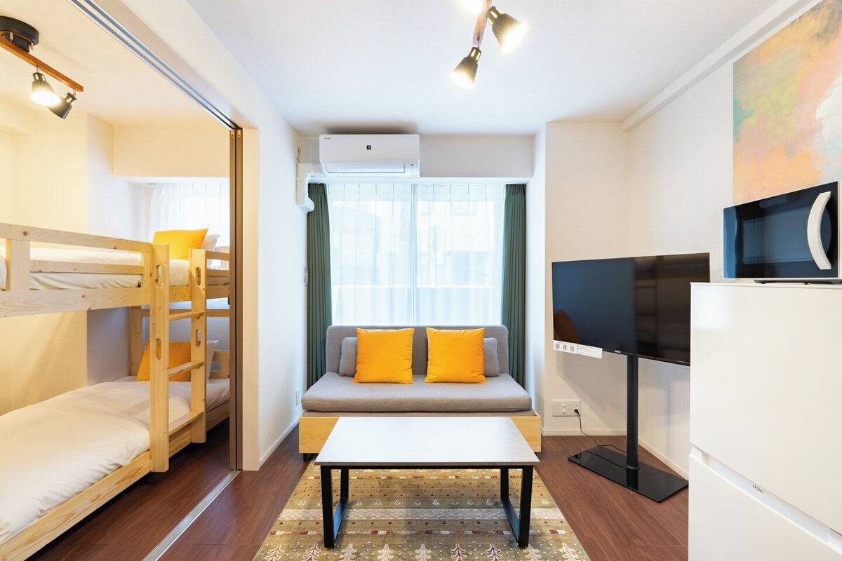 A compact hotel room with a bunk bed, a sofa with cushions, a small table, a flat-screen TV, and a refrigerator.