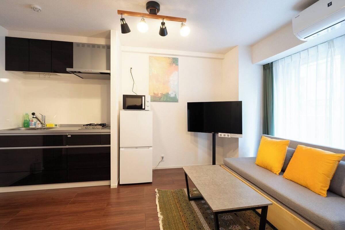 A compact living space with a kitchenette, a flat-screen TV mounted on the wall, a sofa with yellow cushions, and a window with sheer curtains.