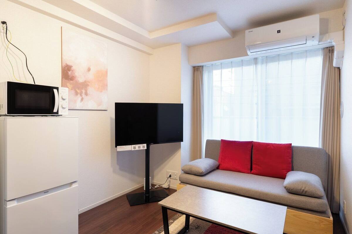 A modern hotel room with a flat-screen TV, a microwave, a mini-fridge, a sofa with red cushions, and a large window with curtains.