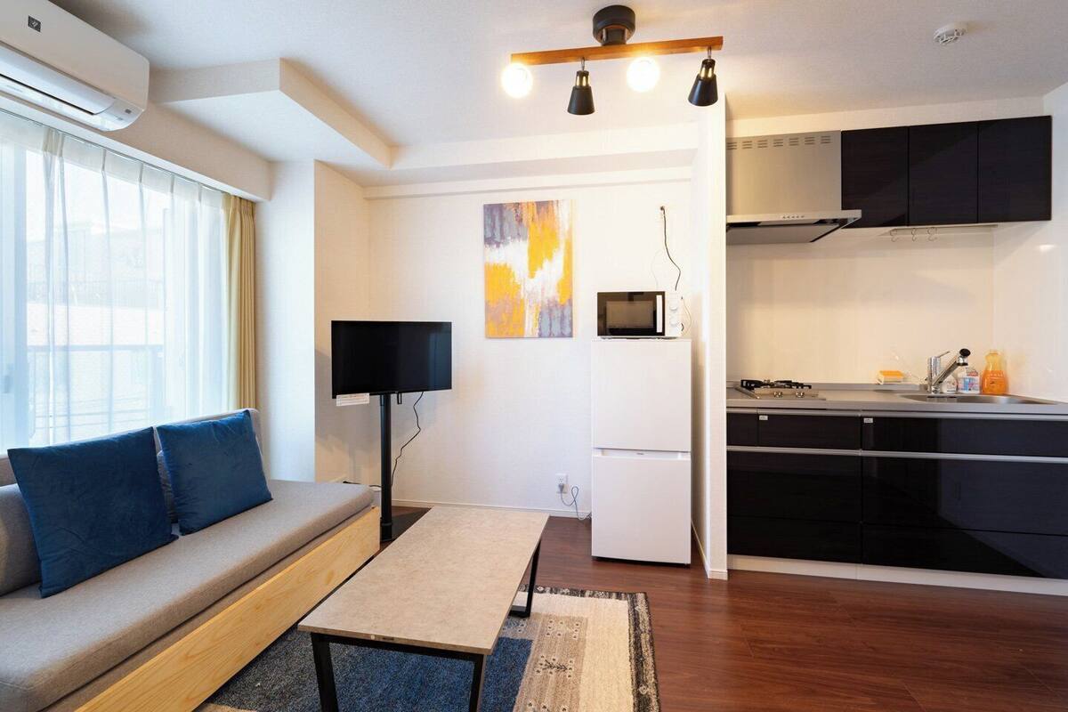 A compact living space with a sofa, a small table, a TV, a kitchen area with a microwave, and a painting on the wall.