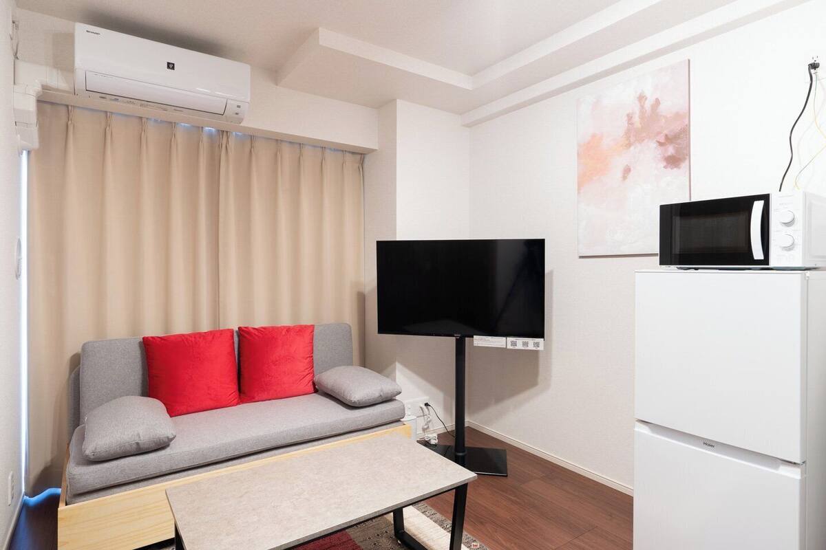 A compact living space with a grey sofa, red cushions, a flat-screen TV, a microwave, and a refrigerator.