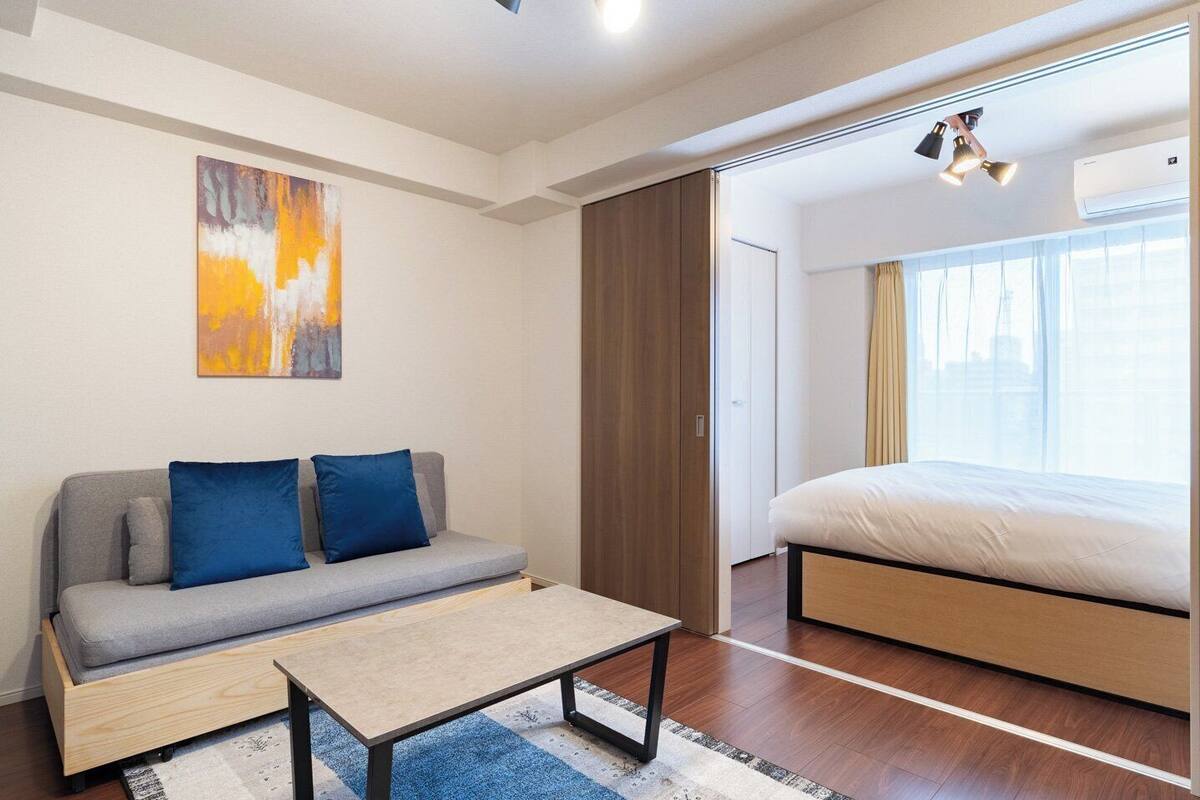 A modern hotel room with a grey sofa, blue cushions, a wooden coffee table, a large bed, and a painting on the wall.