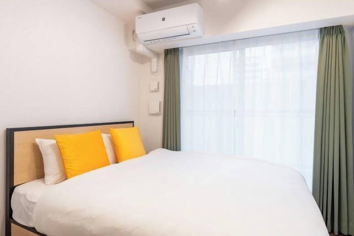 A hotel room with a bed, an air conditioning unit, a window with curtains, and a wall-mounted light switch.