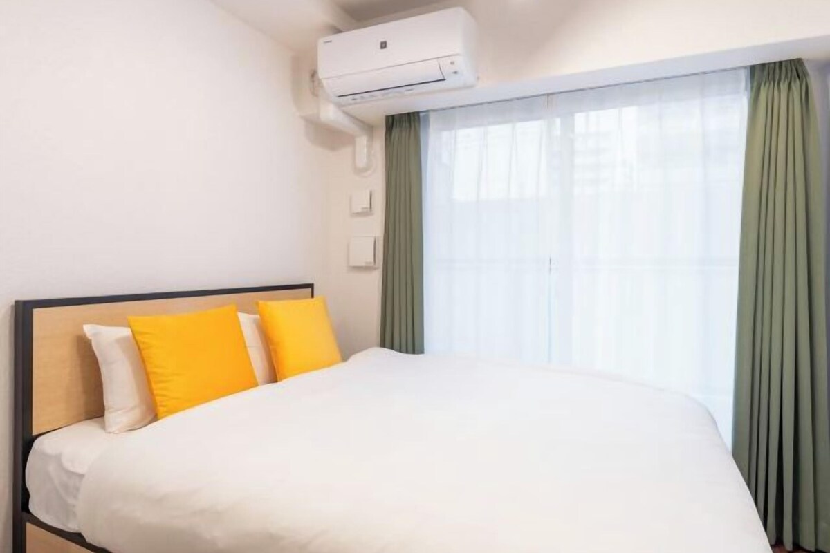 A hotel room with a bed, an air conditioning unit, a window with curtains, and a wall-mounted light switch.