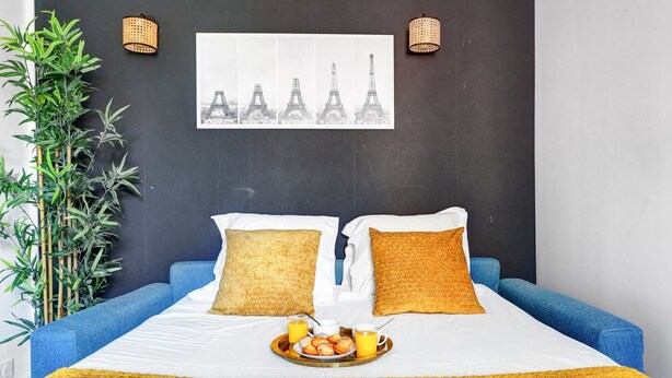 Ein Hotelzimmer mit einem Bett, zwei Kissen, einem Tablett mit Frühstücksangeboten, einem Wandbild von der Eiffelturm und einer Bambuspflanze.