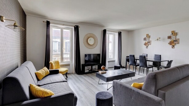 Un salon moderne avec un canapé gris, un fauteuil jaune et une table basse noire. On y trouve également un téléviseur, une table à manger avec des chaises, et un mur agrémenté d’éléments décoratifs.