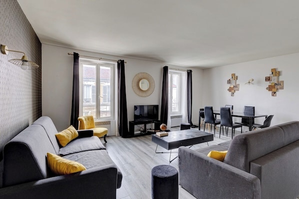Ein modernes Wohnzimmer mit grauem Sofa, gelbem Sessel und einem schwarzen Couchtisch. Ein Fernseher, ein Esstisch mit Stühlen sowie eine Wand mit Dekorationen sind ebenfalls vorhanden.