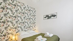 Een netjes opgemaakt bed met een groene deken en twee opgevouwen handdoeken, een nachtkastje met een lamp, en een muur met tropische behang en een ingelijste foto.