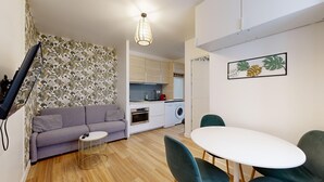 Een compacte leefruimte met een bank, een klein rond tafeltje, een eettafel met stoelen, een keukenhoek met een wasmachine en een aan de muur bevestigde televisie.