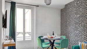 Uma área de refeições com uma mesa redonda posta para dois, cadeiras verdes, uma pequena mesa lateral com uma planta e uma parede estampada.