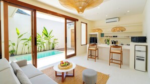 A modern living room with a sofa, a coffee table with fruit, a kitchen with a fridge, and a pool view. - Villa Joba by Pertama Stay (Canggu)