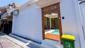 A modern building with a white exterior, a wooden door with intricate carvings, and a small pool area visible through the doorway. - Villa Joba by Pertama Stay (Canggu)