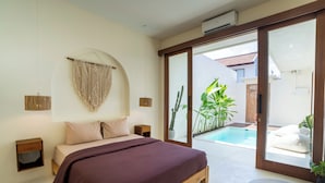 A bedroom with a bed, bedside tables, a hanging macrame wall decor, and a sliding glass door leading to a pool area with plants. - Villa Joba by Pertama Stay (Canggu)