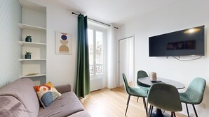 Ein modernes Wohnzimmer mit einer Couch, einem Esstisch mit Stühlen, einem an der Wand montierten Fernseher und einem Fenster mit Vorhängen.