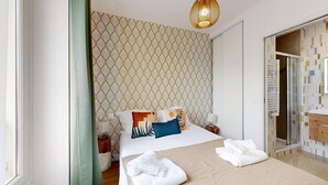 Ein Hotelzimmer mit strukturiertem Wandpapier, einem Bett mit vielen Kissen, einem Nachttisch mit Lampe sowie einem Badezimmer mit Duschkabine aus Glas und einem Holzschrank.