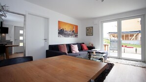 Un salon moderne avec une table à manger en bois, un canapé noir agrémenté de coussins colorés, un tableau représentant un paysage urbain et une grande porte-fenêtre coulissante donnant sur un espace extérieur.