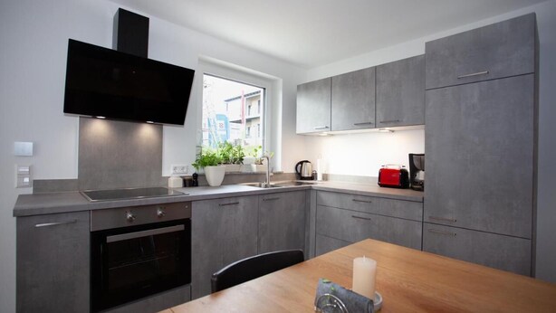 Une cuisine moderne avec des meubles gris, un four encastré, un téléviseur à écran plat et une fenêtre laissant entrer la lumière naturelle.