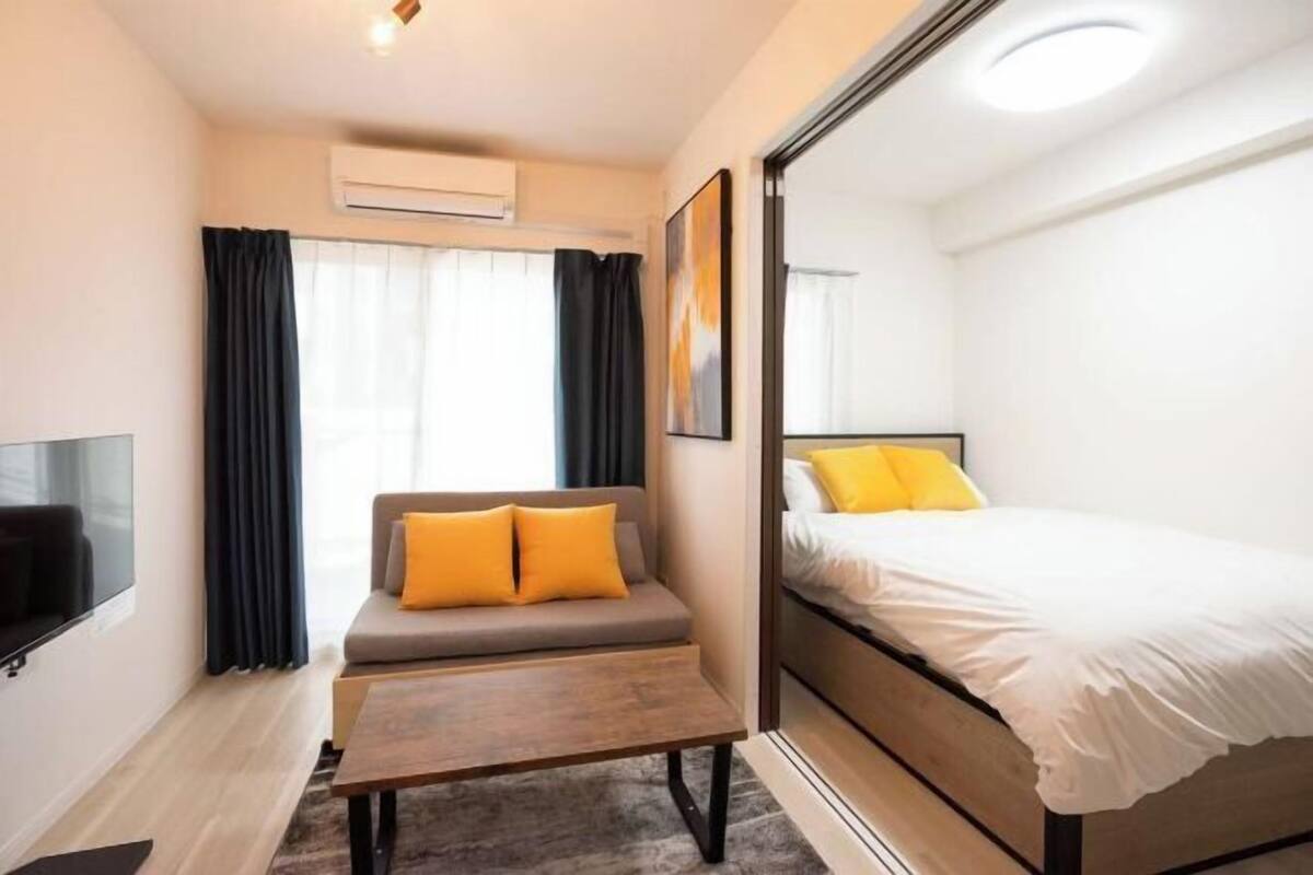 A compact hotel room with a sofa, a bed, a flat-screen TV, and a window with curtains.