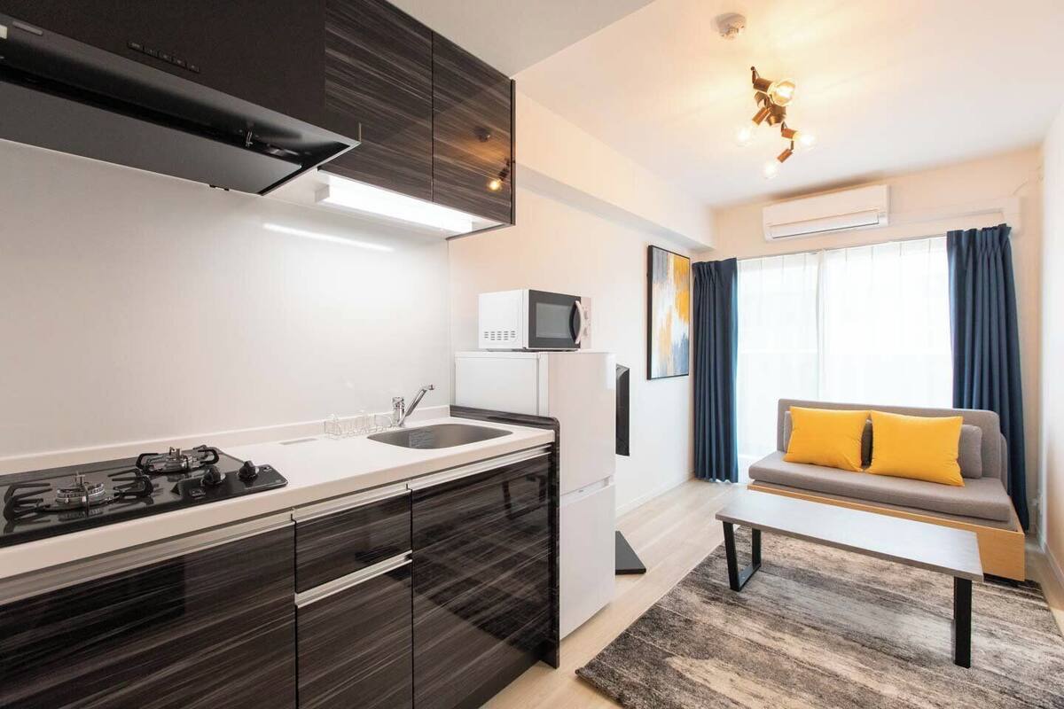 A modern kitchen with a gas stove, white cabinets, and a microwave. There is a small dining area with a table and a sofa with yellow cushions.