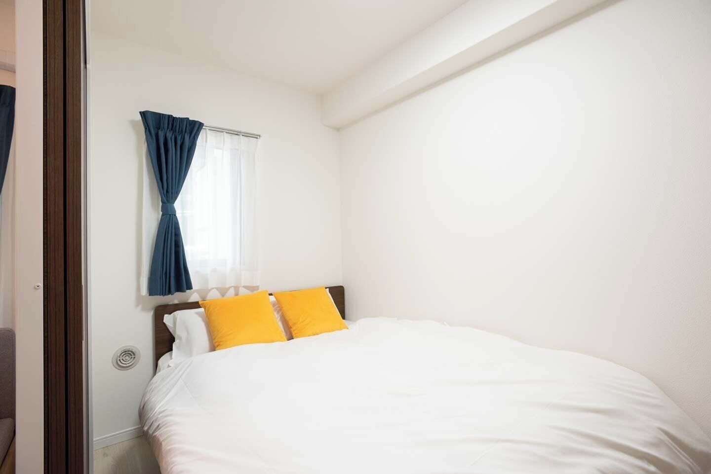 Une chambre à coucher avec un lit, des murs blancs, une fenêtre avec des rideaux bleus et une tête de lit en bois.