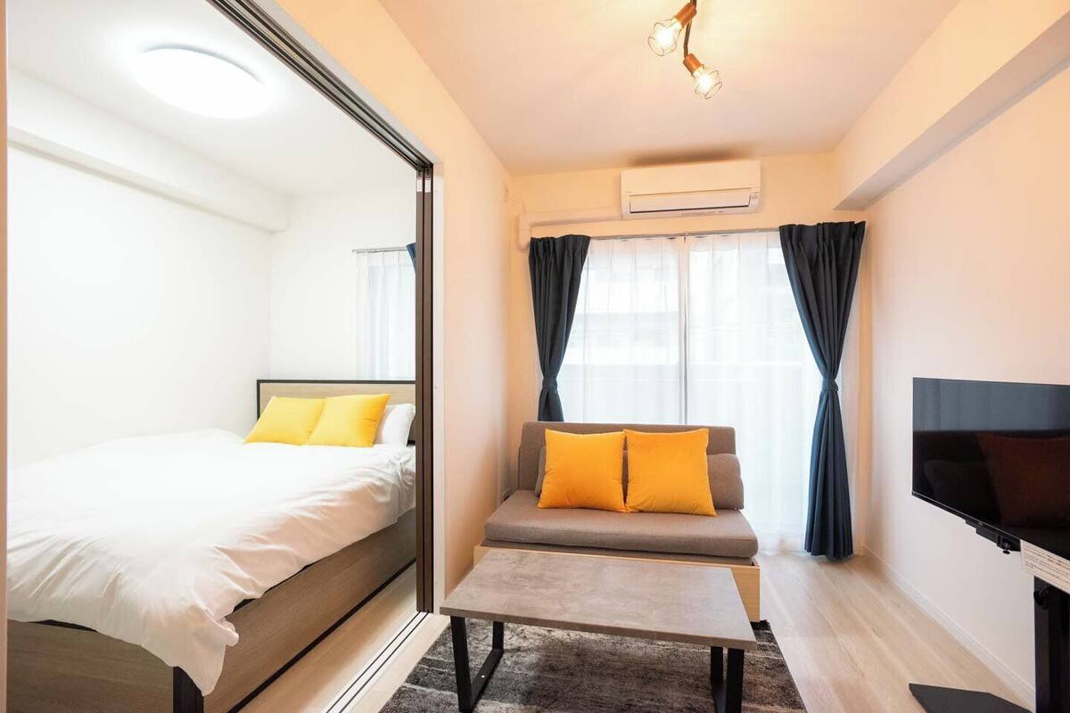 A modern hotel room with a bed, a sofa with yellow cushions, a small table, a flat-screen TV, and an air conditioning unit.