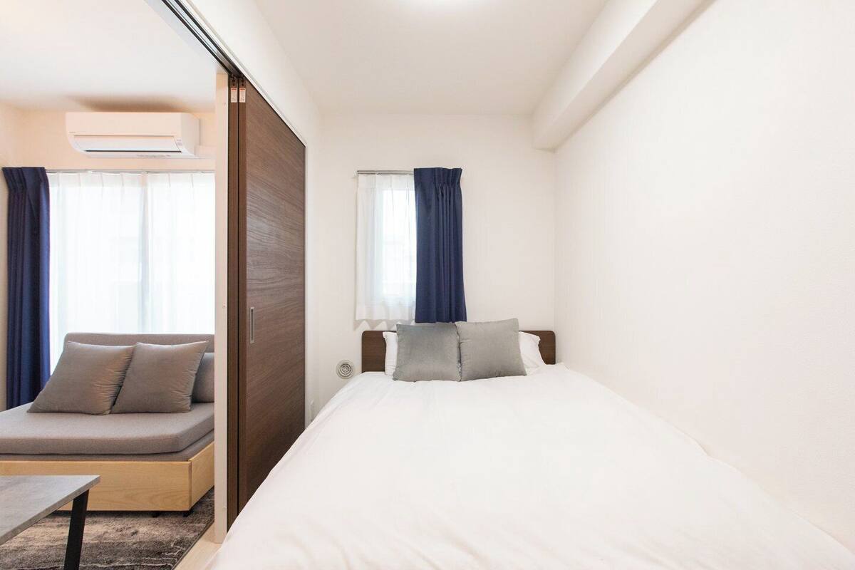 A hotel room with a bed, a sofa, a window with curtains, and an air conditioning unit.