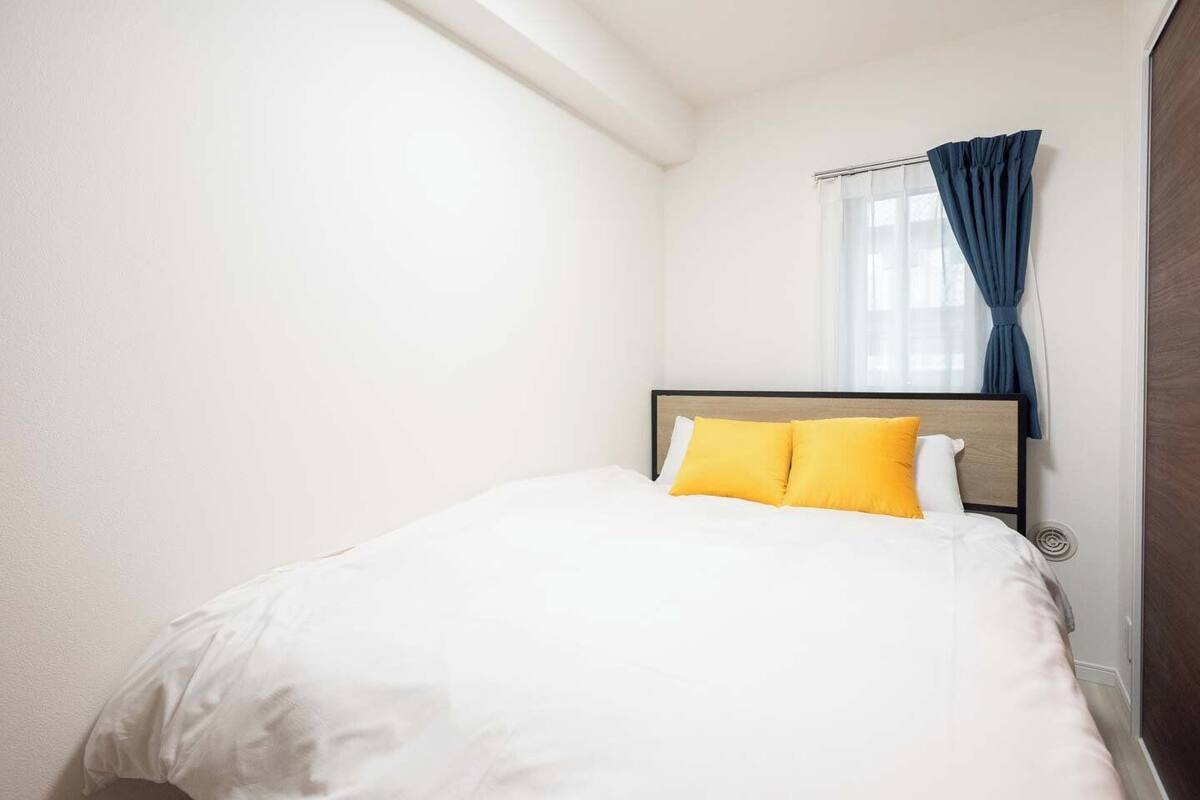 A neatly made bed with white bedding and two yellow pillows, a window with blue curtains, and a wooden headboard.