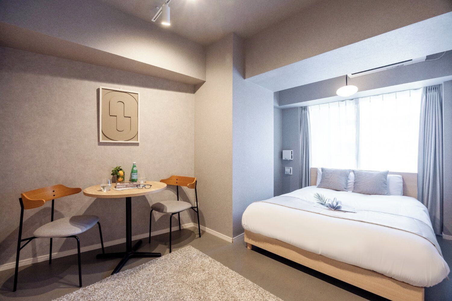 A hotel room with a bed, a desk with two chairs, a small table with a plant and a bottle, and a window with curtains.