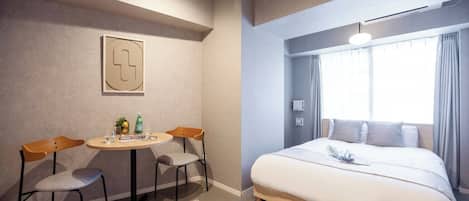 A hotel room with a bed, a desk with two chairs, a small table with a plant and a bottle, and a window with curtains.