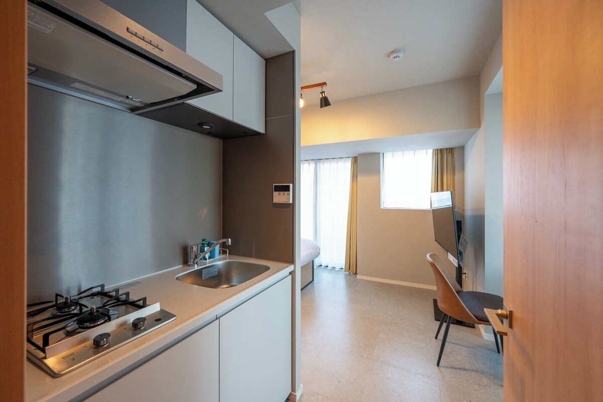 A modern kitchen with a gas stove, a sink, and a built-in oven. There is a dining area with a table and chairs, and a TV mounted on the wall.