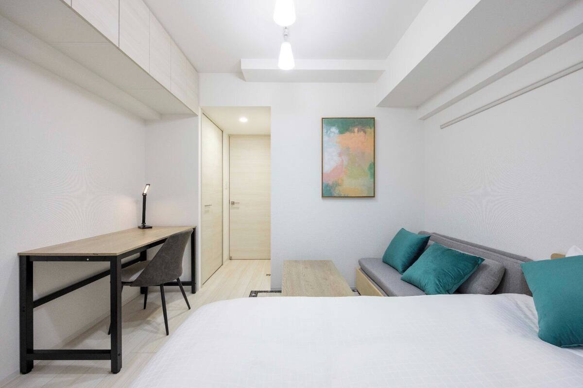 Ein modernes Schlafzimmer mit Bett, Schreibtisch und Stuhl. Eine Tür führt in einen anderen Raum, und ein Gemälde hängt an der Wand.
