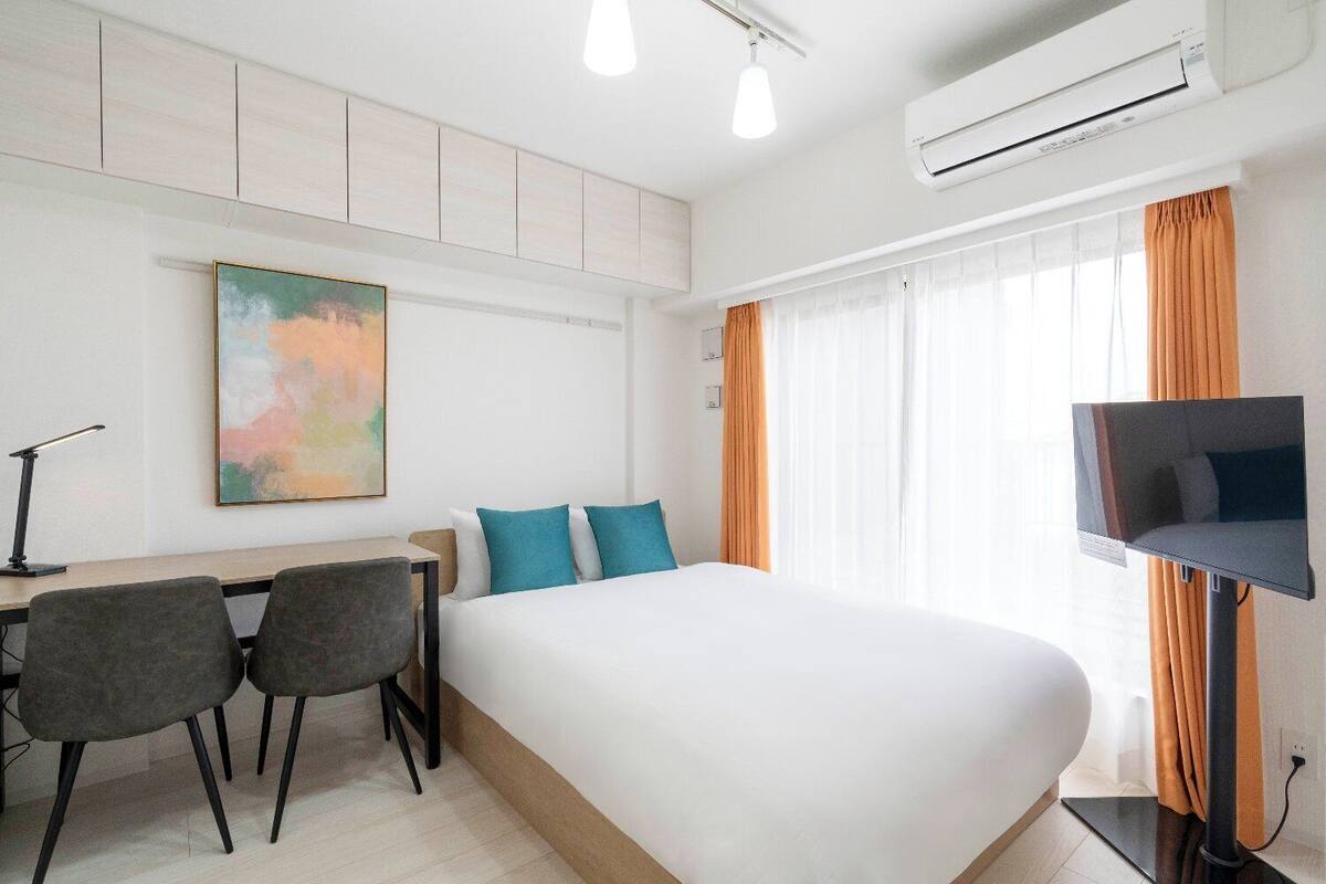 Ein Hotelzimmer mit Bett, Schreibtisch, zwei Stühlen, einer Schreibtischlampe, einer Klimaanlage und einem Fernseher.