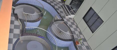 Un espacio exterior escalonado con asientos circulares y suelo a cuadros, rodeado por un edificio con ventanas y una puerta.