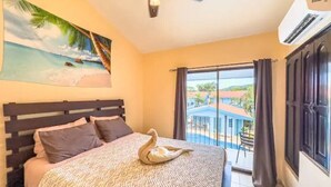 Una camera da letto con un letto, una ventola a soffitto, un quadro raffigurante una spiaggia e vista su una piscina e degli edifici.
