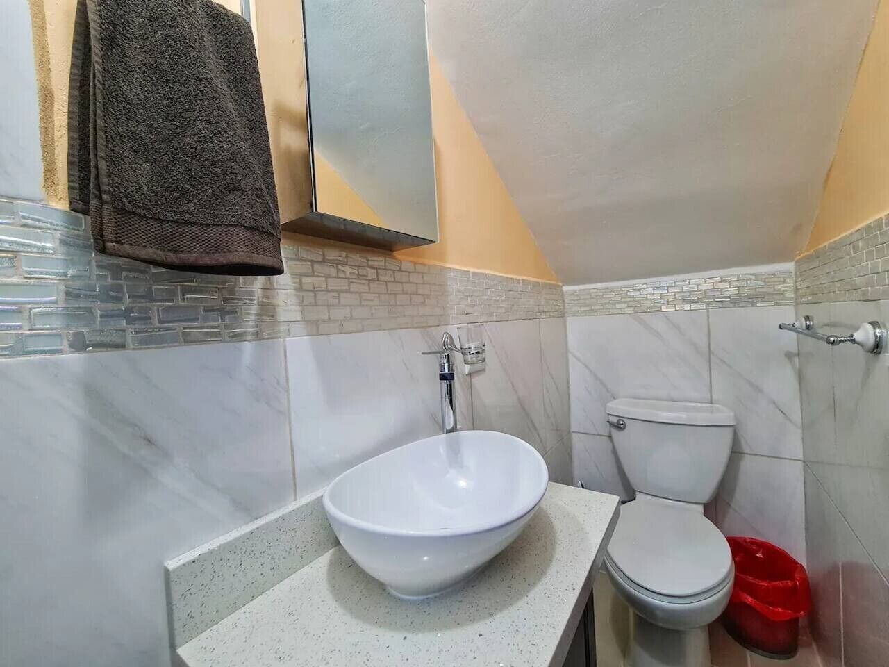 Un baño con un lavamanos blanco tipo recipiente, un inodoro y un cubo de basura rojo.