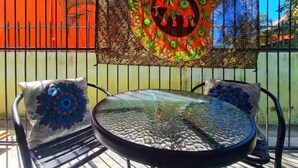 Uma mesa de vidro redonda com cadeiras de metal, uma almofada estampada e uma tapeçaria colorida numa parede.