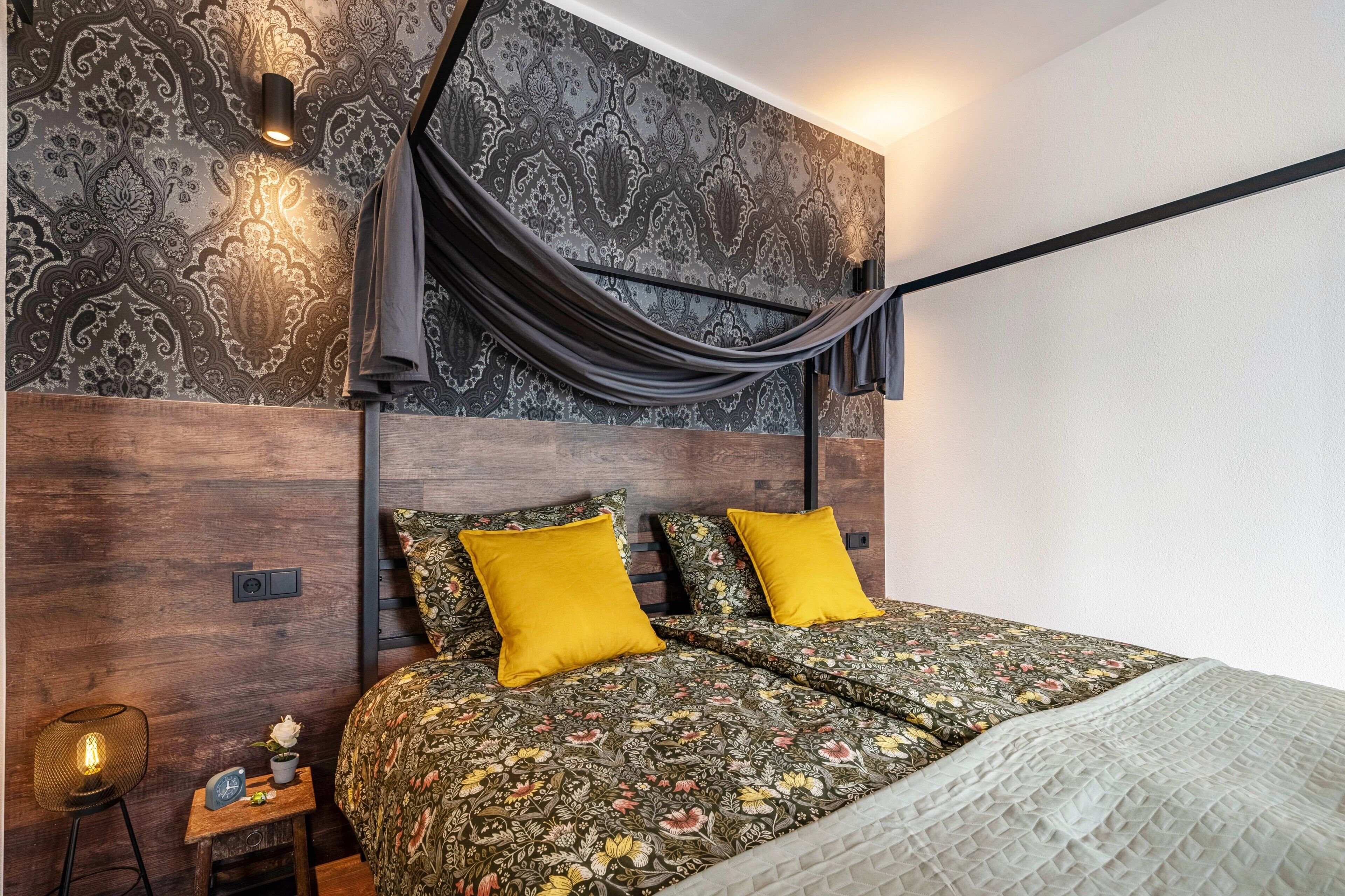 Ein Schlafzimmer mit ornamentalem Wandpapier, einem Bett mit floraler Bettwäsche, einem schwarzen Kopfteil und einem Nachttisch mit Lampe.